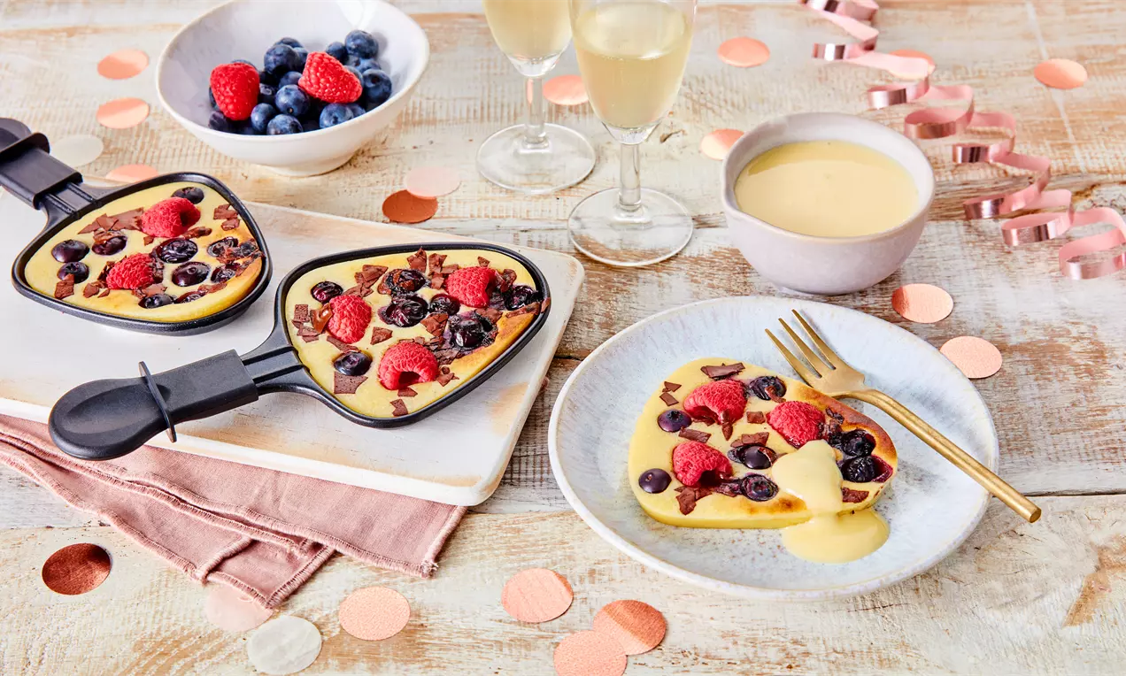 Geschmolzener süßer Teig in Raclettepfännchen mit frischen Himbeeren und Blaubeeren, goldener Gabel auf cremeweißem Teller, dekorativ angerichtet