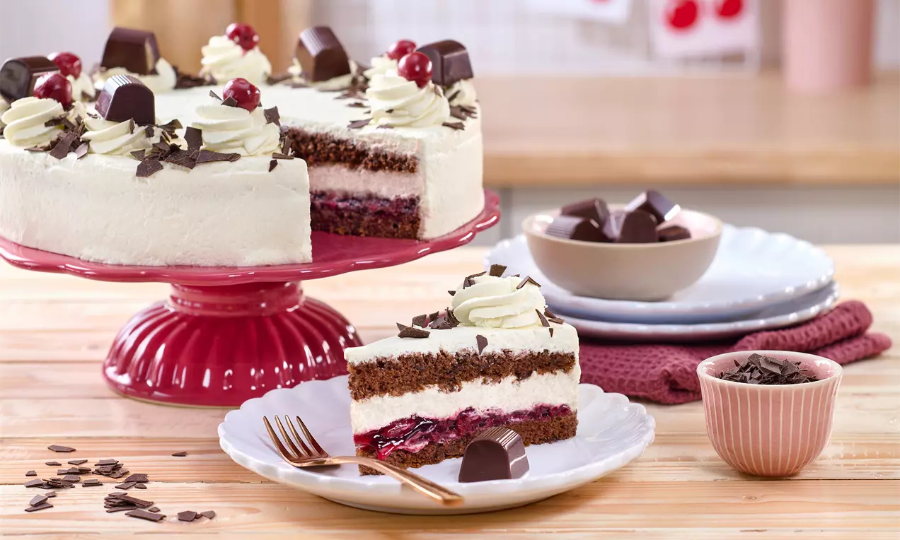 Mon-Chéri-Torte auf einer roten Tortenplatte; helle Creme, Schokobiskuit, Kirschfüllung und Sahnetuffs, dekoriert mit Schokoraspeln, Kirschen und Pralinen; ein Stück liegt auf einem Teller im Vordergr