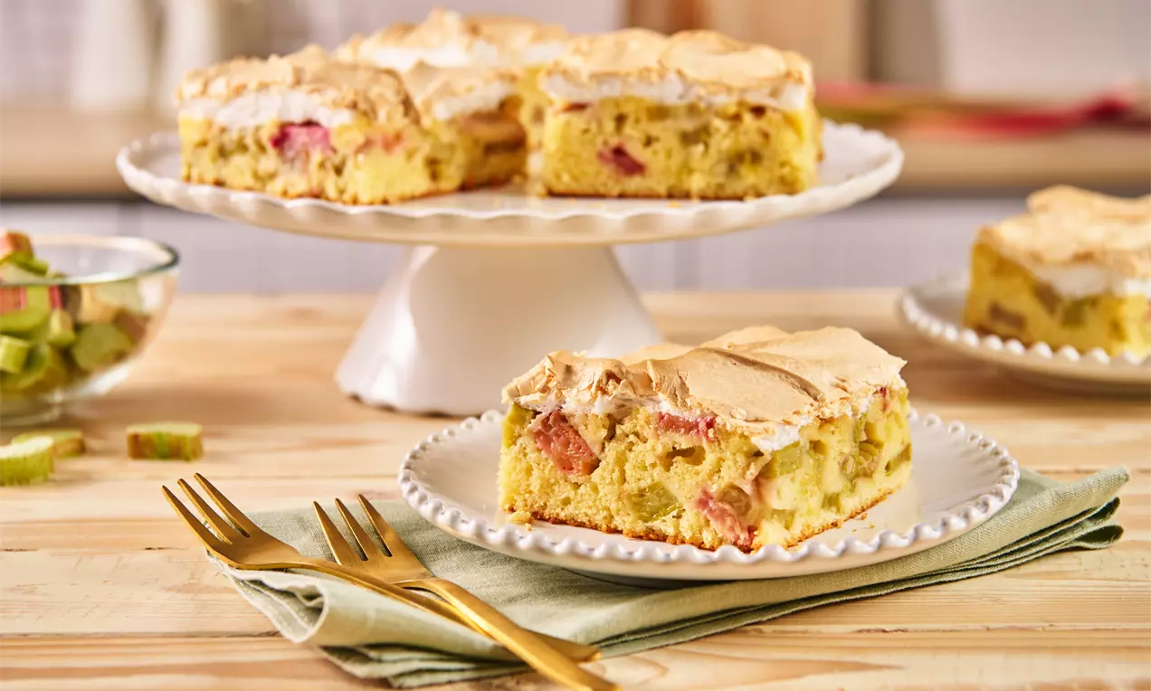 Ein goldgelber Rhabarberkuchen mit luftigem Baiser auf einer weißen Tortenplatte, dazu ein Stück auf einem Teller mit goldenen Gabeln