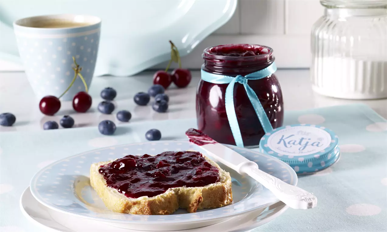 Ein Glas mit tiefroter Konfitüre, verziert mit blauem Schleifenband, daneben ein mit Konfitüre bestrichener Brotscheiben auf Teller