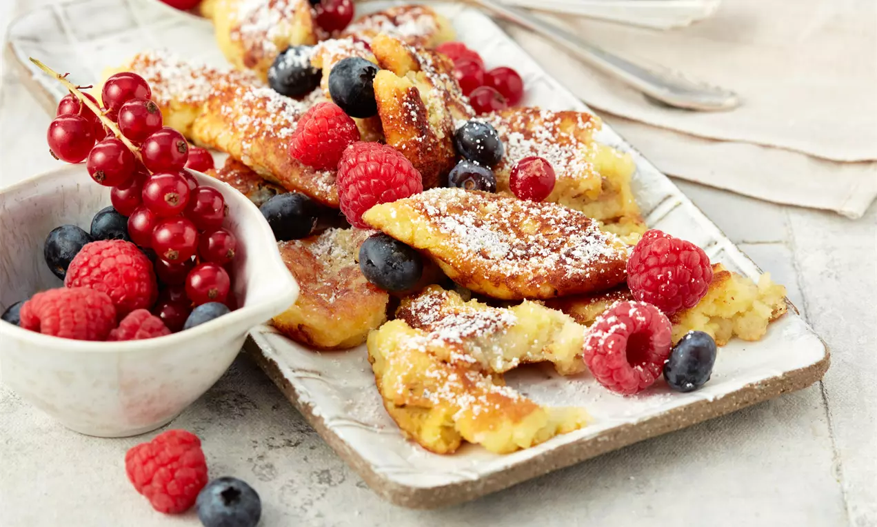 Goldgelber, grob zerrissener Kaiserschmarrn mit Puderzucker bestäubt und frischen Himbeeren, Blaubeeren und roten Johannisbeeren garniert