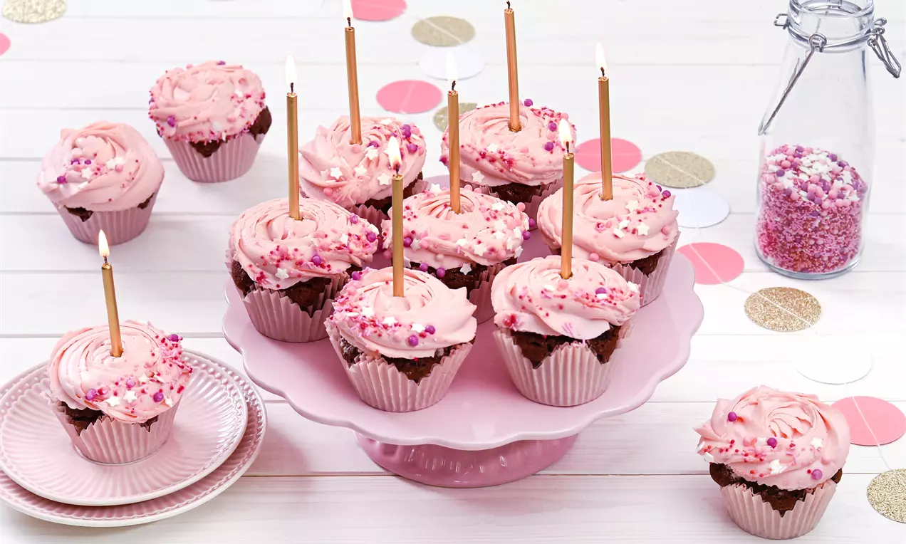 Rosa Cupcakes mit cremigem Frosting und Streuseln, verziert mit goldenen Kerzen, auf rosa Tortenplatte und hellem Holztisch arrangiert