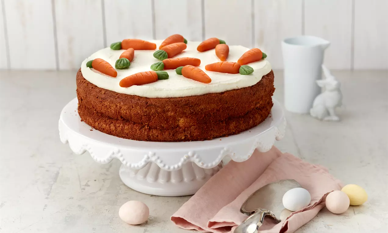 Rüblitorte mit hellbraunem Boden und weißer Creme, dekoriert mit Marzipankarotten, auf einer weißen Tortenplatte mit rosa Stofftuch