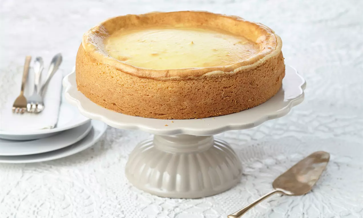 Goldbrauner Käsekuchen mit glatter, leicht glänzender Oberfläche auf einer weißen Tortenplatte, arrangiert auf hellem Spitzendeckchen