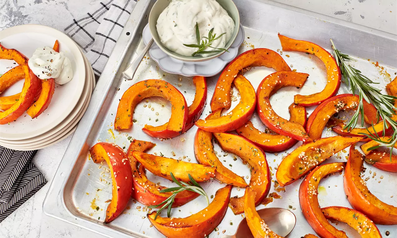 Geröstete orange Kürbisspalten mit goldbraunem Rand auf einem Backblech, garniert mit Kräutern, neben cremigem Dip in einer Schale