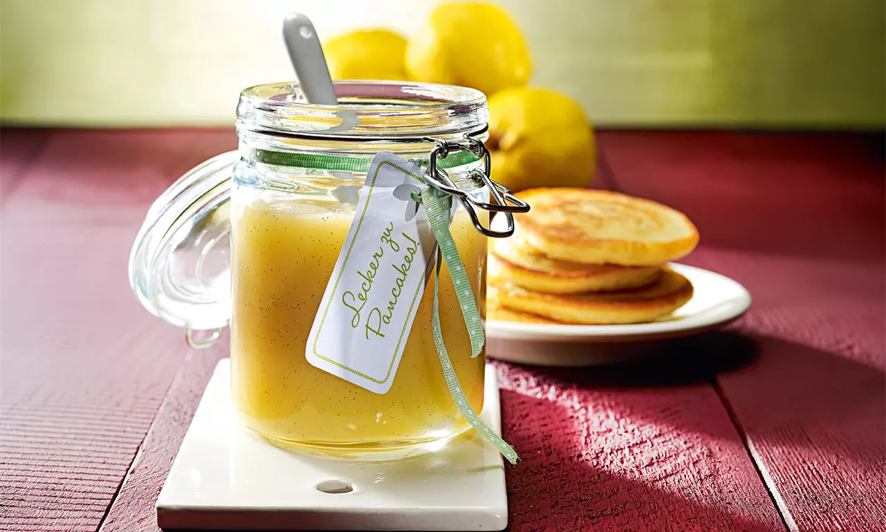 Glas mit hellgelbem Quitten-Apfelmus, verziert mit grünem Band und Etikett, daneben ein Teller mit goldbraunen Pancakes