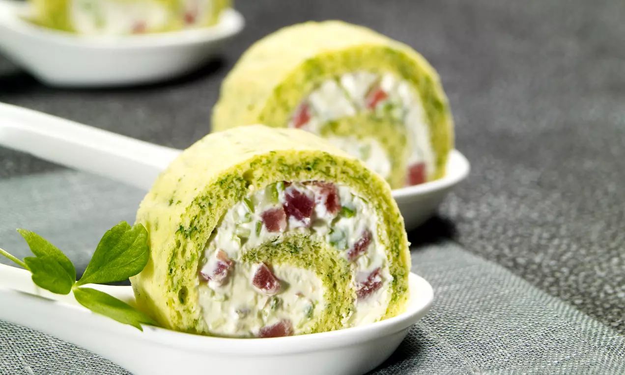 Scheiben einer grünlichen Schinken-Frischkäse-Rolle mit weißer Creme, roten Stücken und Kräutern auf weißen Porzellanlöffeln angerichtet