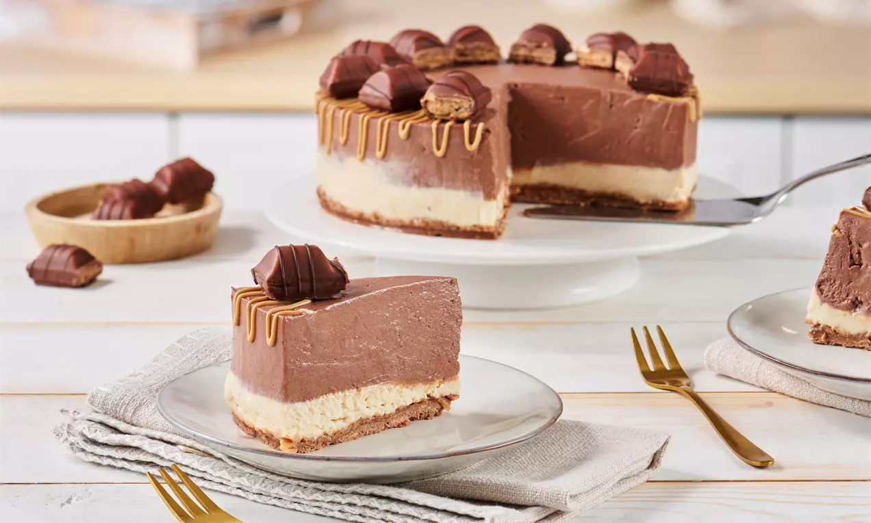 Cremige Torte mit Schichten aus heller und dunkler Füllung auf knusprigem Boden, dekoriert mit Schokoriegelstücken und Karamellzartstreifen