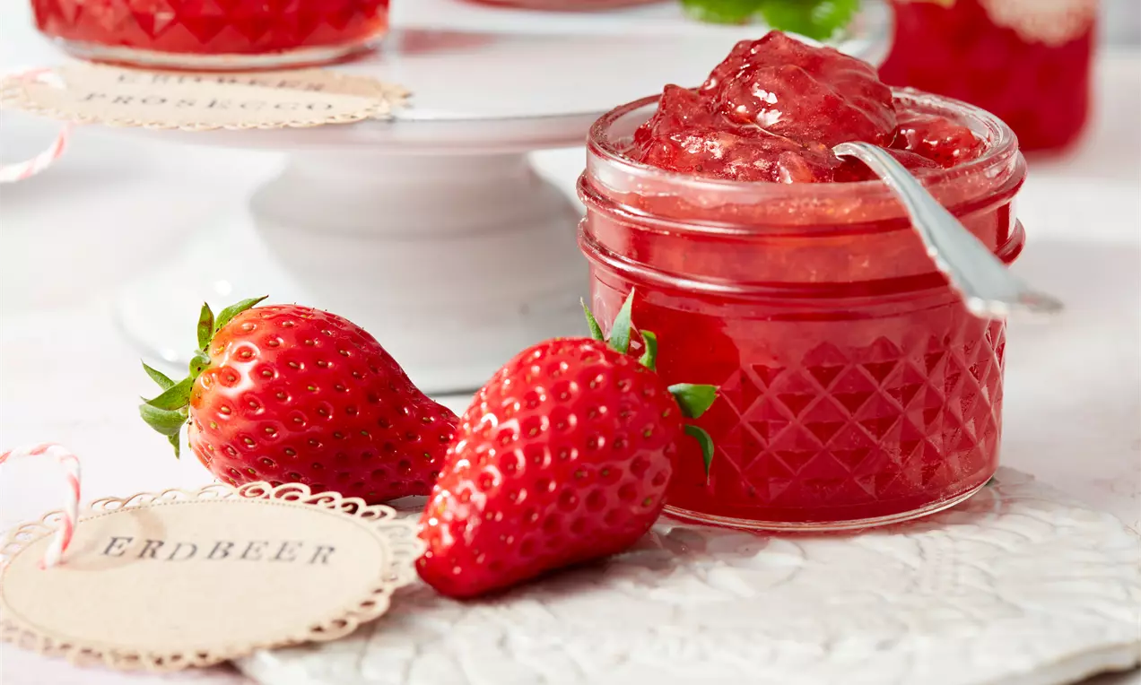 Erdbeer-Prosecco-Konfitüre in einem kleinen Glas mit Rautenmuster, gefüllt mit roter, glänzender Konfitüre neben frischen Erdbeeren und Etikett