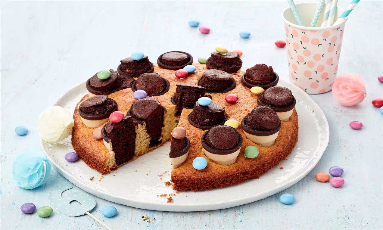 Runder, goldbrauner Kinder-Partykuchen auf weißer Platte, dekoriert mit Schokoladenstückchen, Sahnehauben und bunten Schokolinsen