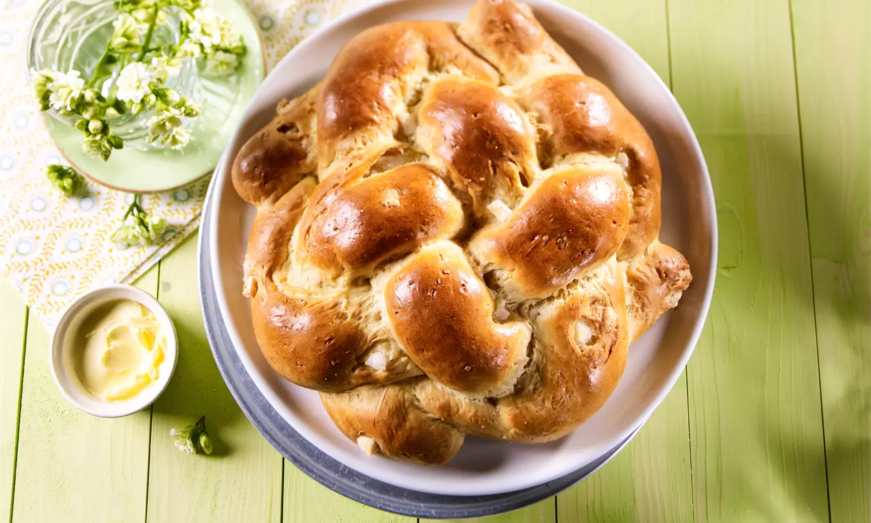 Goldbrauner süßer Flechtknoten mit glänzender Oberfläche auf einem weißen Teller, daneben Butter in einer Schale und Blumen auf hellem Holztisch