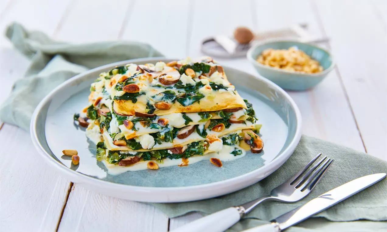 Spinat-Schafkäse-Lasagne mit geschichteten gelben Nudelplatten, grünem Spinat, weißer Sauce und gerösteten Nüssen auf Keramikteller