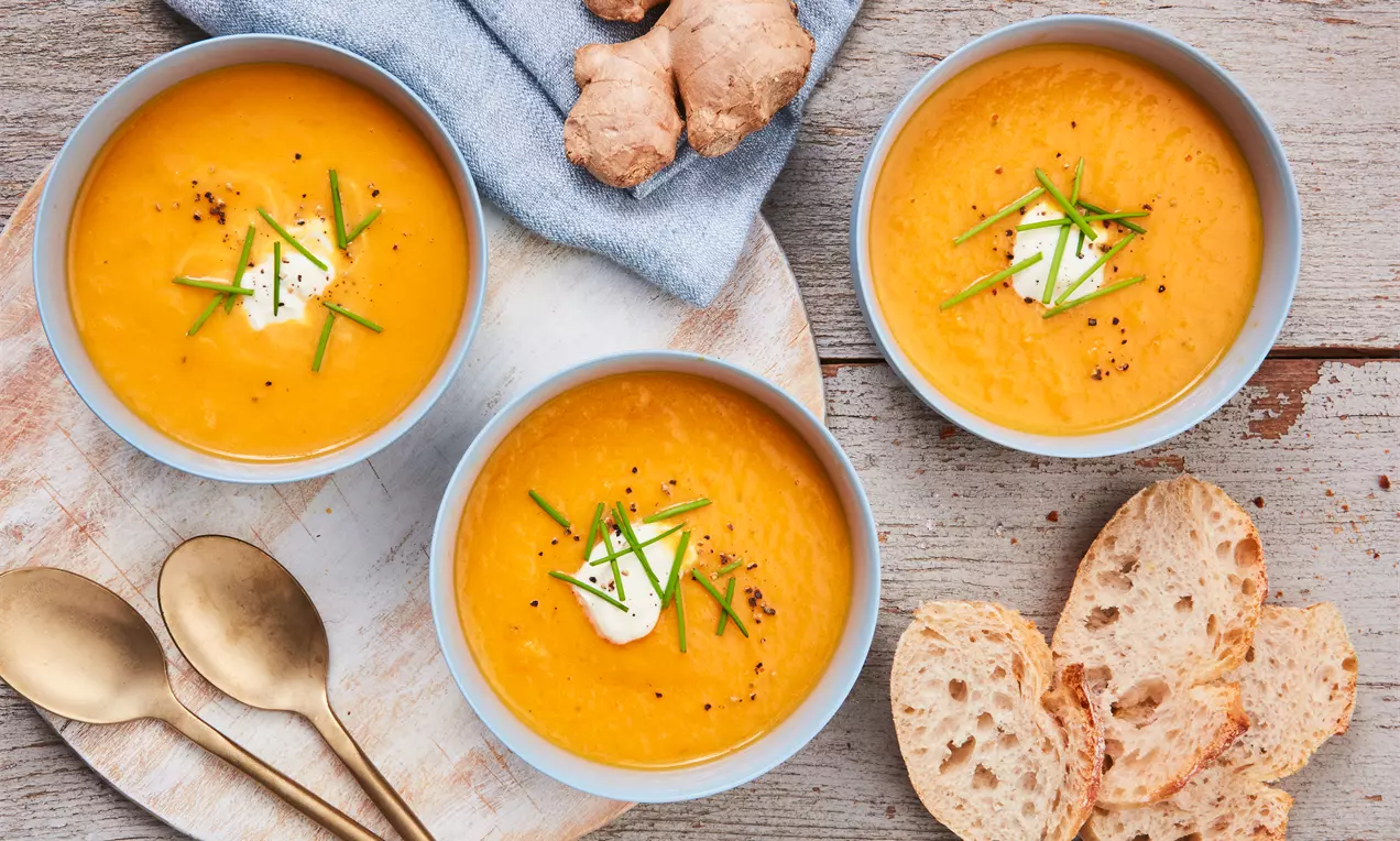 Drei Schalen mit samtig orangefarbener Möhren-Ingwer-Suppe, garniert mit einem Klecks Creme, Schnittlauch und Pfeffer, dazu Brotscheiben