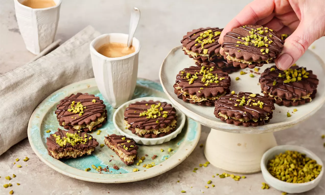Dunkelbraune Doppelkekse mit Schokoglasur und gehackten Pistazien auf gelblich-grünen Tellern, daneben Tassen mit Kaffee und Pistazienschale