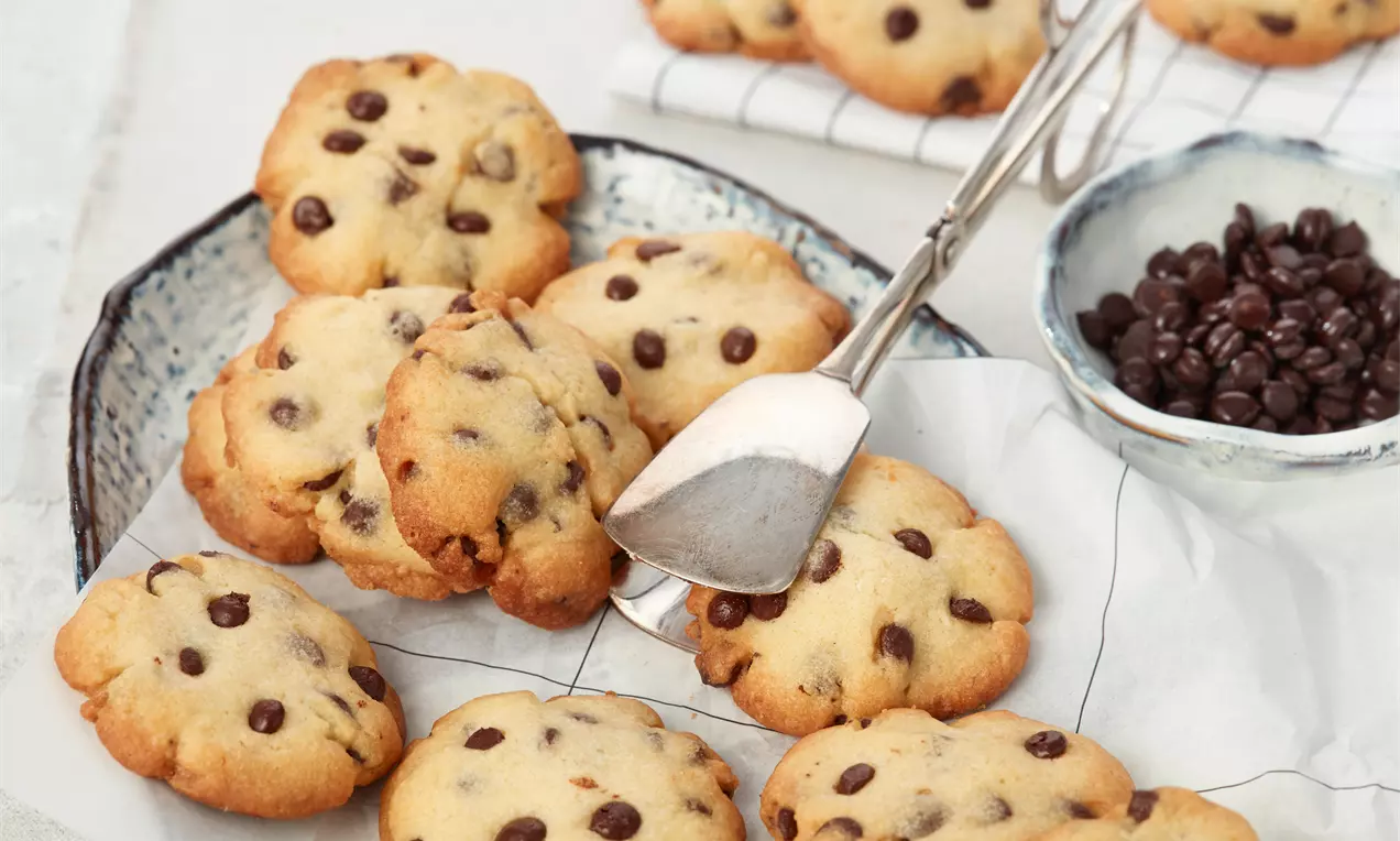 Goldbraune Chocolate-Chip-Cookies mit dunklen Schokostückchen auf weißem Papier in einer rustikalen Schale, daneben Schokotropfen in kleiner Schale