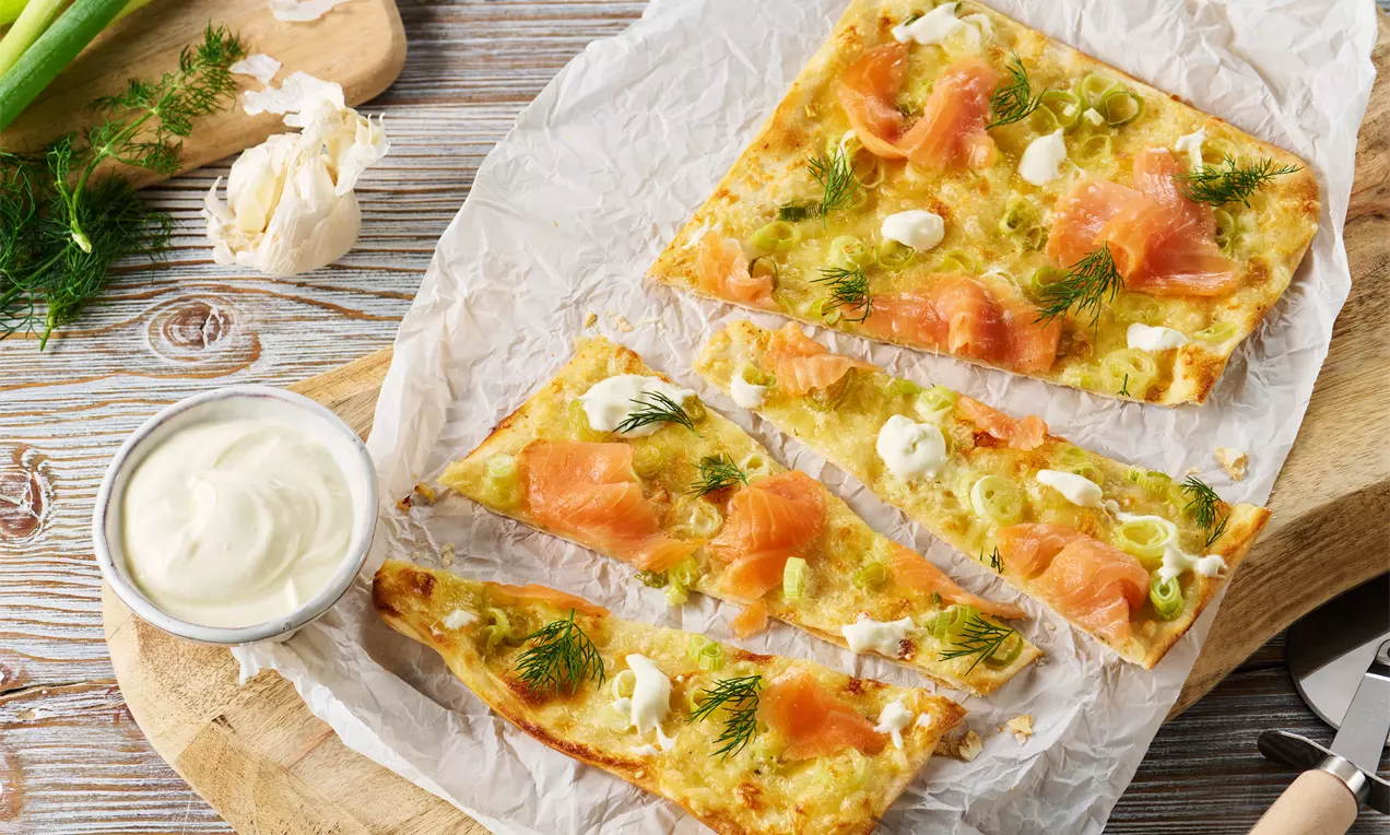Flammkuchen mit knusprigem Teig, Lachsscheiben, Frühlingszwiebeln, Dill und Klecksen von Crème fraîche auf Pergamentpapier serviert