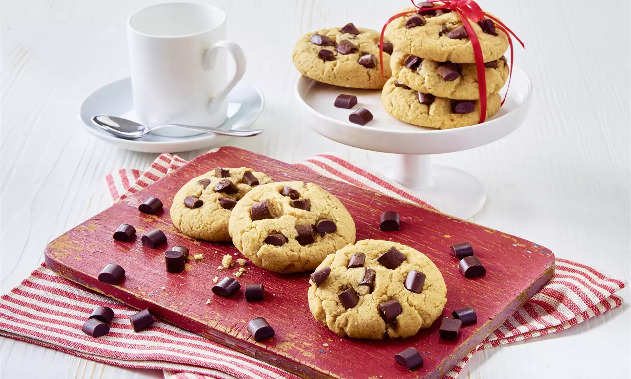 Goldbraune Cookies mit großen, dunklen Schokoladenstückchen auf rotem Holzbrett und Streifentuch, daneben eine Tasse und ein Teller voller Cookies