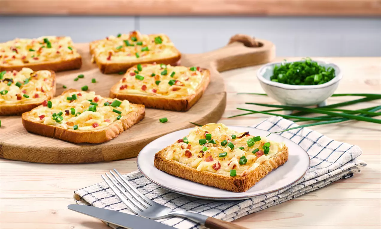 Flammkuchen-Toast mit goldbrauner Kruste, belegt mit cremiger Schicht, Speckwürfeln und grünem Schnittlauch auf rustikalem Holzbrett