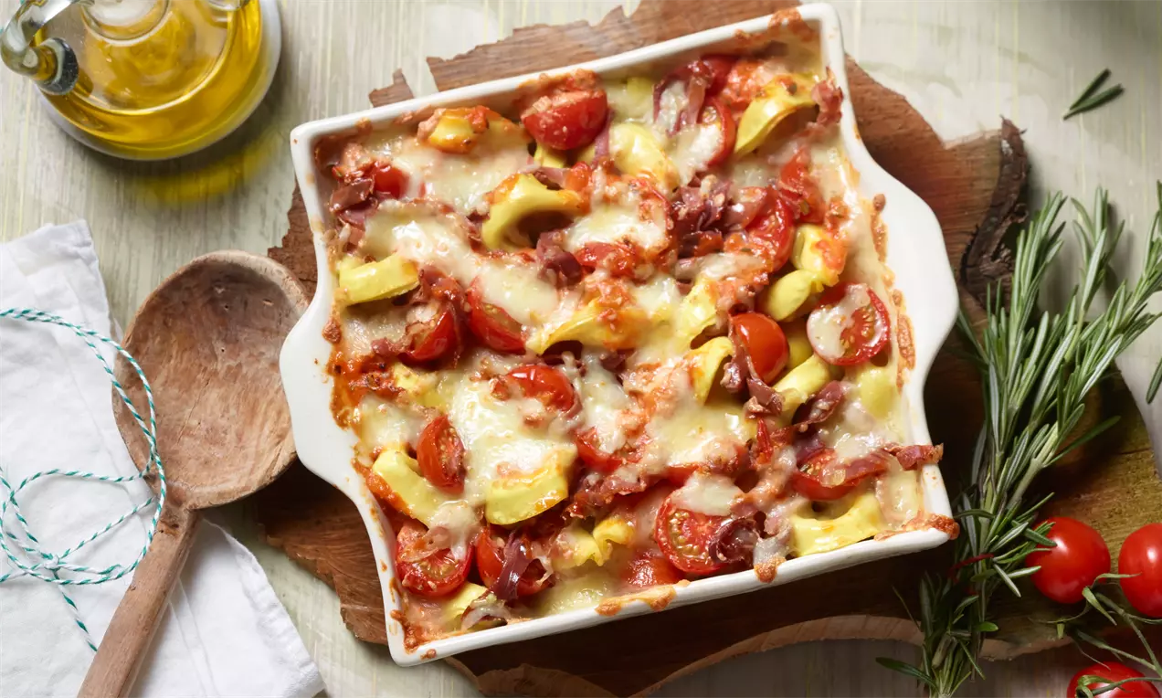 Tortelloni-Auflauf mit Parmaschinken, geschmolzenem Käse, leuchtend roten Kirschtomaten und grünen Rosmarinzweigen in goldbrauner Kruste