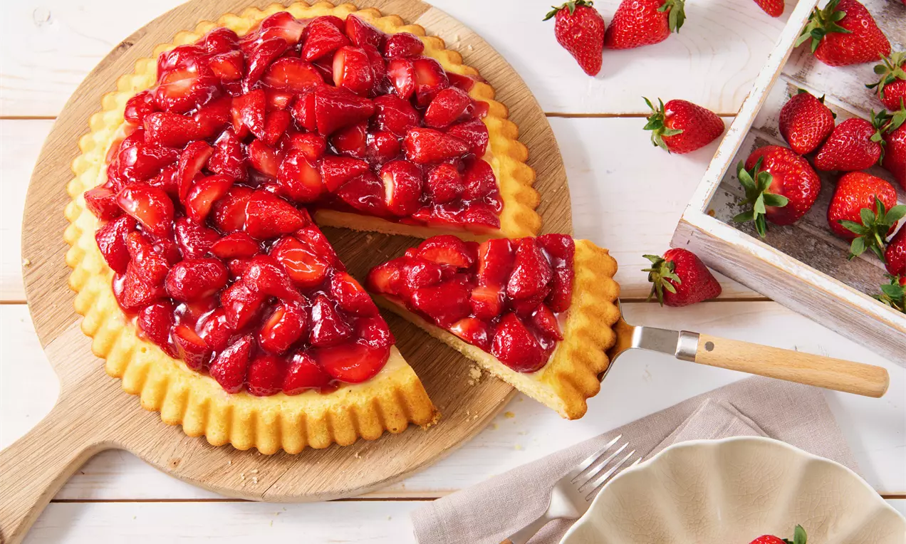Goldgelber Rührteigkuchen belegt mit glänzenden roten Erdbeeren, ein Stück wird herausgeschnitten, serviert auf Holzbrett mit Deko-Erdbeeren