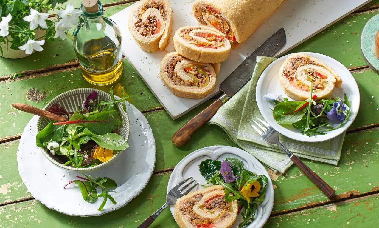 Eine goldgelbe, aufgeschnittene Teigrolle mit fleischhaltiger Füllung, angerichtet auf Tellern mit frischem, grünem Salat und essbaren Blüten