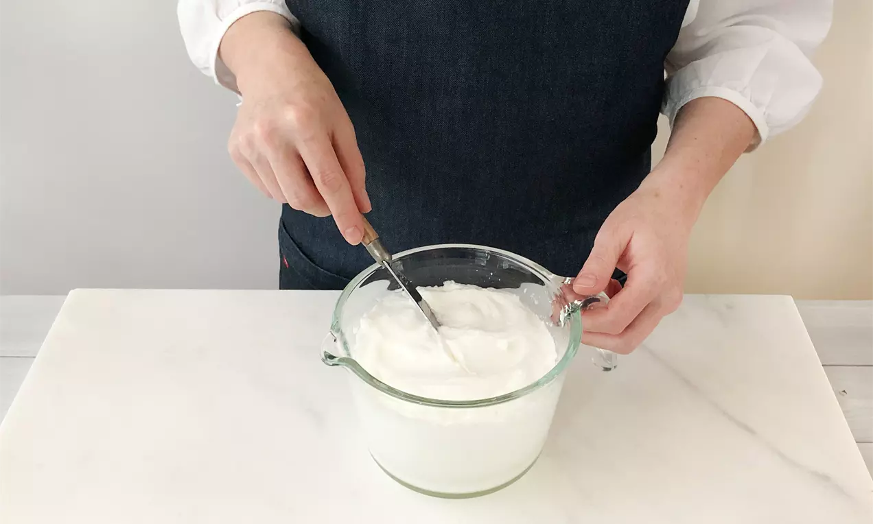 Schneeweißes, cremiges Eiweiß in einer transparenten Glasschüssel, auf einer weißen Arbeitsfläche von Händen mit Schneebesen bearbeitet