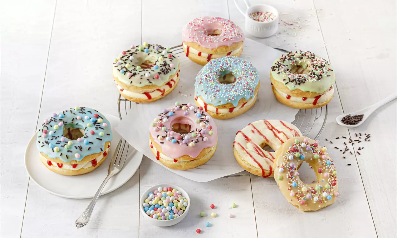 Donuts mit glänzenden Zuckerglasuren in Pastellfarben, bunten Streuseln und Puddingfüllung auf weißem Teller und Holztisch drapiert