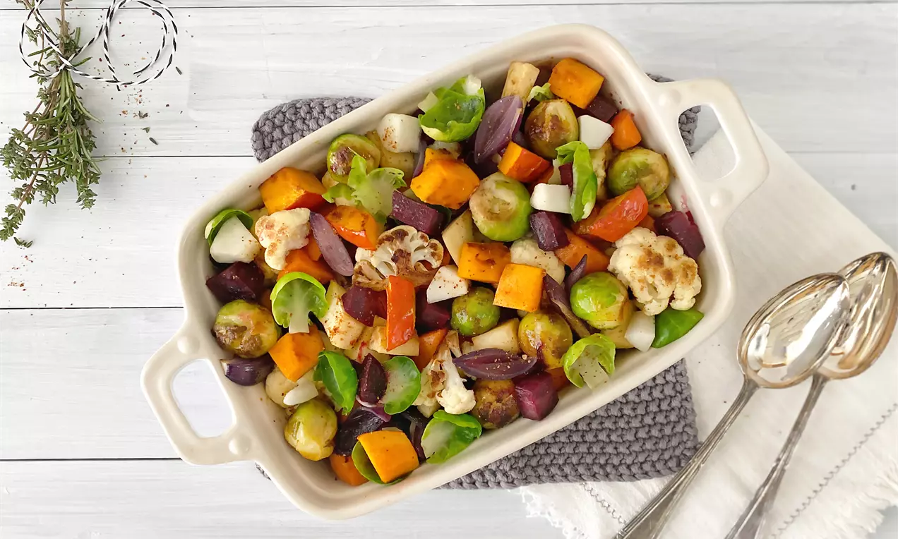 Buntes Ofengemüse mit goldbraunen Röstspuren aus Rosenkohl, Süßkartoffeln, roten Zwiebeln und Blumenkohl in heller Auflaufform auf Stoffunterlage
