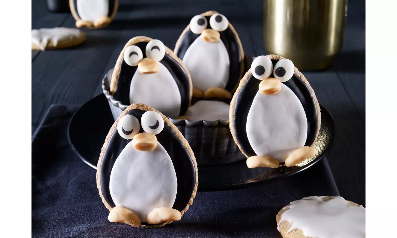 Pinguin-Plätzchen mit weißem Bauch und schwarzen Flügeln aus Zuckerguss, dekoriert mit Augen und orangefarbenem Schnabel auf dunklem Tuch