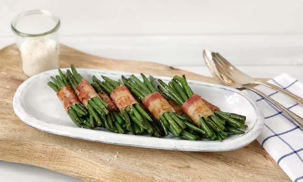 Schmale Bohnenbündel umwickelt mit knusprigem, goldbraunem Speck auf länglicher weißer Platte, daneben Besteck und weiß-blaues Tuch