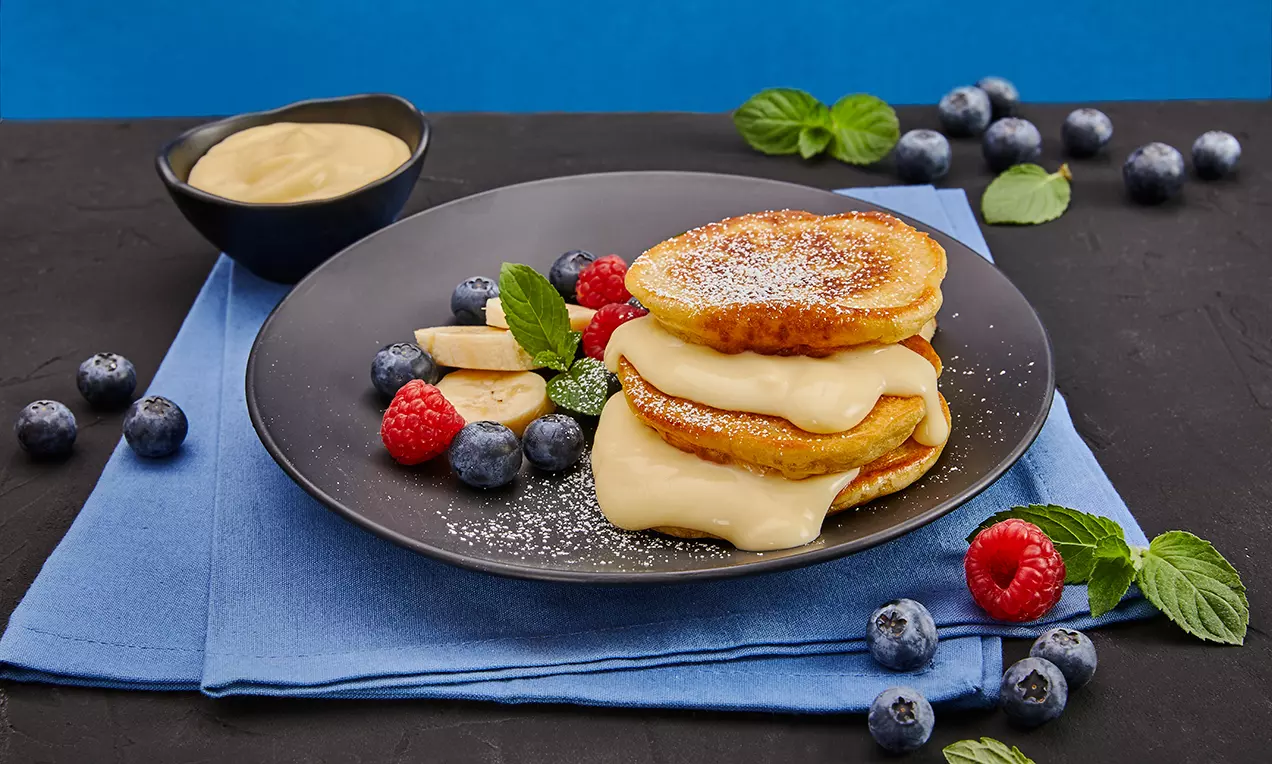 Gefüllte Protein-Pancakes mit Bananenscheiben und Vanillepuddingfüllung, goldbraun auf weißem Teller mit Beeren und Ahornsirup