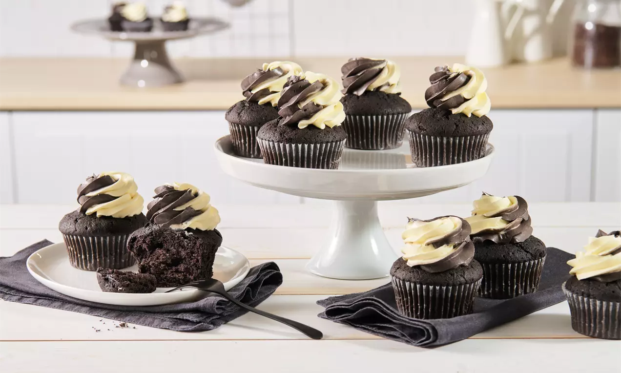 Schokoladencupcakes mit weißer und dunkler Cremehaube auf weißer Etagere und Teller, arrangiert mit Stoffservietten im hellen Küchenambiente