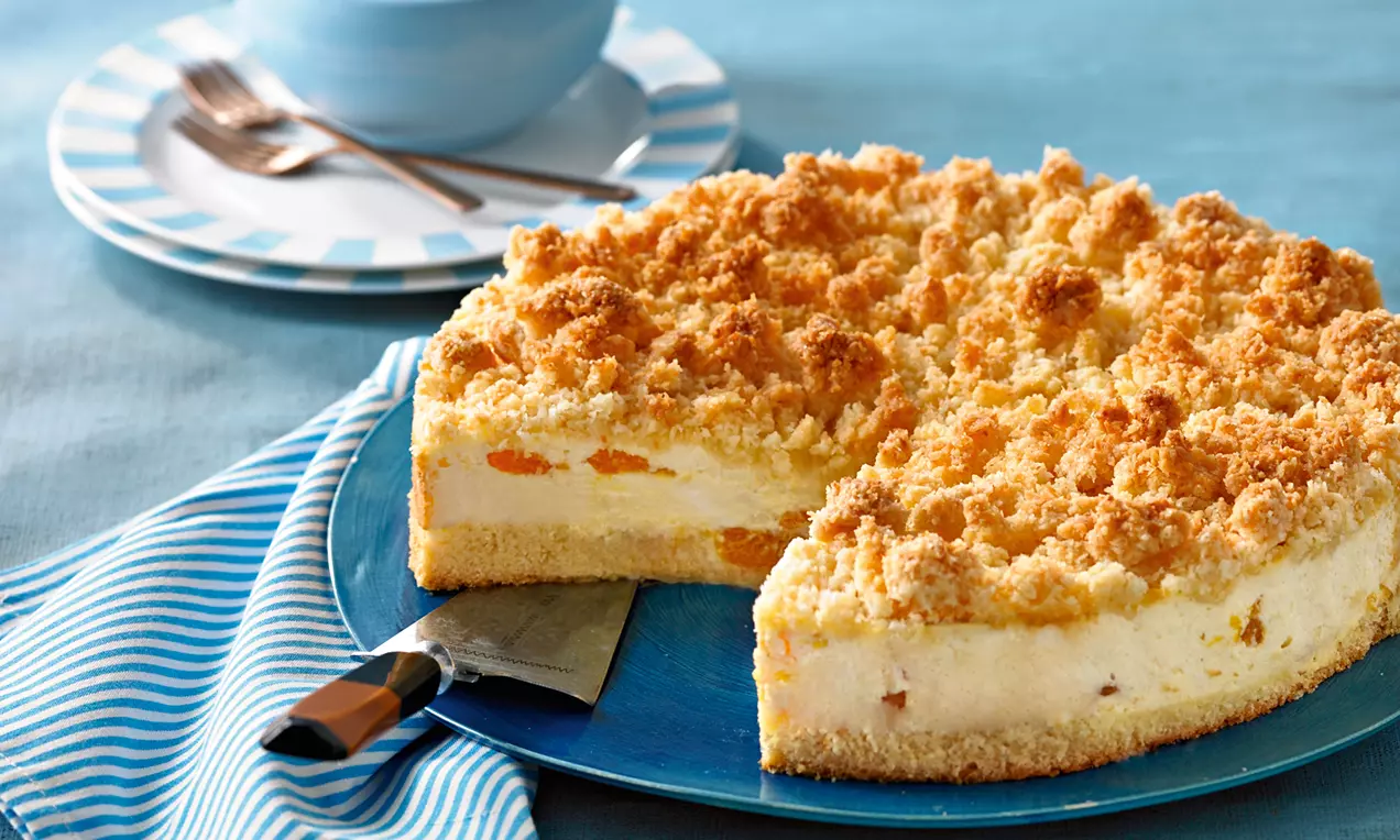 Kokosstreusel-Käsekuchen mit goldbrauner Kruste, cremiger Füllung und Fruchtstückchen auf blauem Teller, daneben gestreiftes Tuch und Besteck