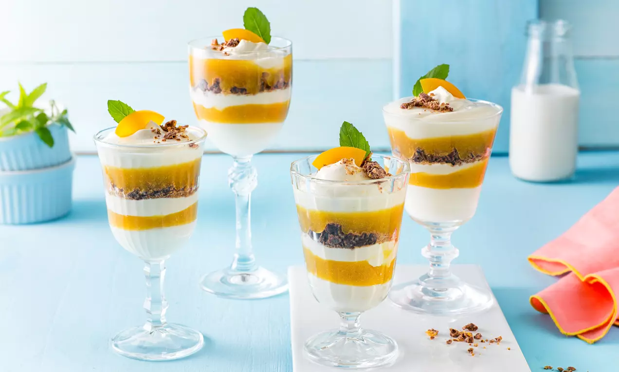 Schichtdesserts in hohen Gläsern, abwechselnd weiße Creme, goldgelbes Fruchtpüree und dunkle Streusel, mit Minzblättern und Aprikosenspalten garniert