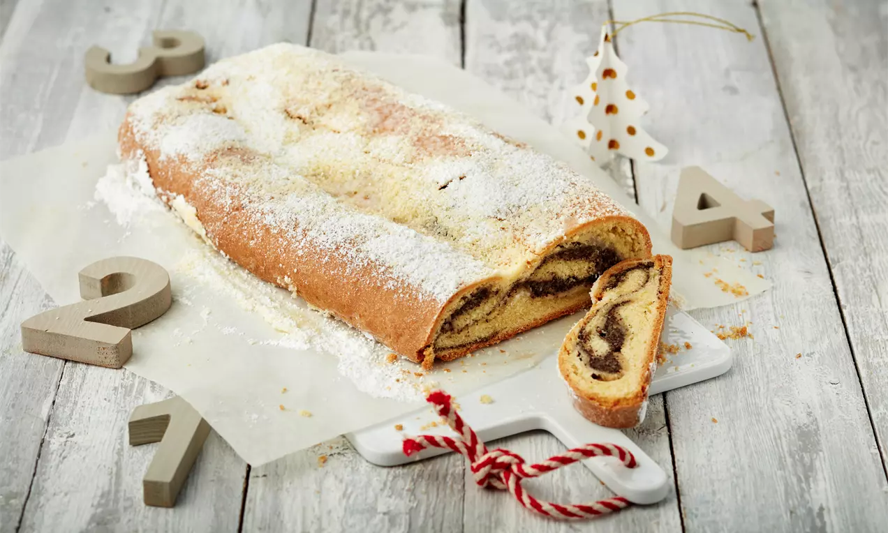 Quarkstollen mit goldbrauner Kruste, Puderzucker bestäubt, eine Scheibe mit dunkler Mohn-Marzipan-Füllung auf weißem Holzbrett mit Deko