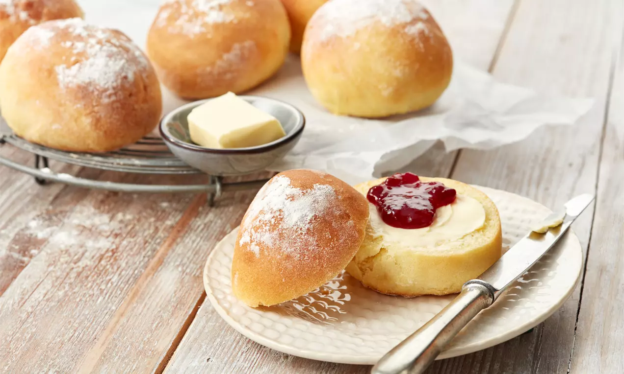 Goldbraune Brötchen mit weicher, fluffiger Textur, bestäubt mit Mehl, eines halbiert mit Butter und roter Marmelade auf einem Teller
