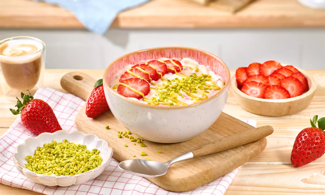 Skyr-Pudding in weißer Schale mit frischen Erdbeerscheiben und gehackten Pistazien, auf Holzbrett mit Löffel, daneben Pistazien und Erdbeeren