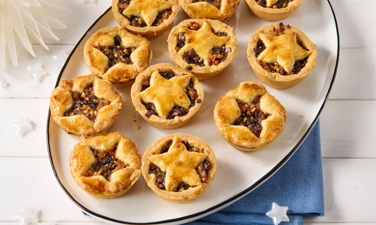 Goldbraune, sternförmig verzierte Mince Pies mit knusprigem Teig und dunkler Fruchtfüllung auf einem ovalen Teller mit blauer Serviette drapiert