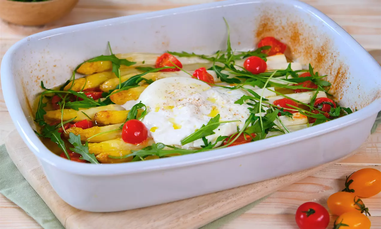 Goldbrauner Spargel im weißer Auflaufform mit saftigen Tomaten, geschmolzener Burrata und frischem Rucola auf einem Holzbrett drapiert