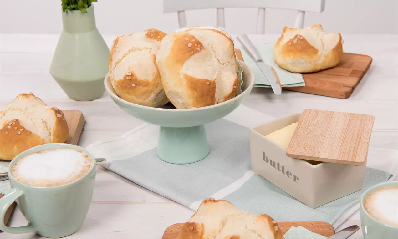 Goldbraune Laugenbrötchen mit salziger Kruste auf Holzbrett und mintgrüner Tortenplatte, umgeben von Cappuccino und Butterbox in heller Tischszene