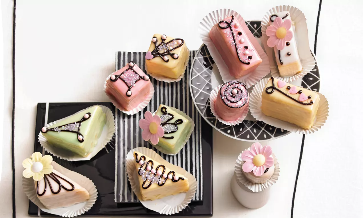 Petits Fours in pastellfarbenem Marzipan mit feiner Verzierung, auf Papierförmchen und Tellern angerichtet, teils mit Blütendekoration