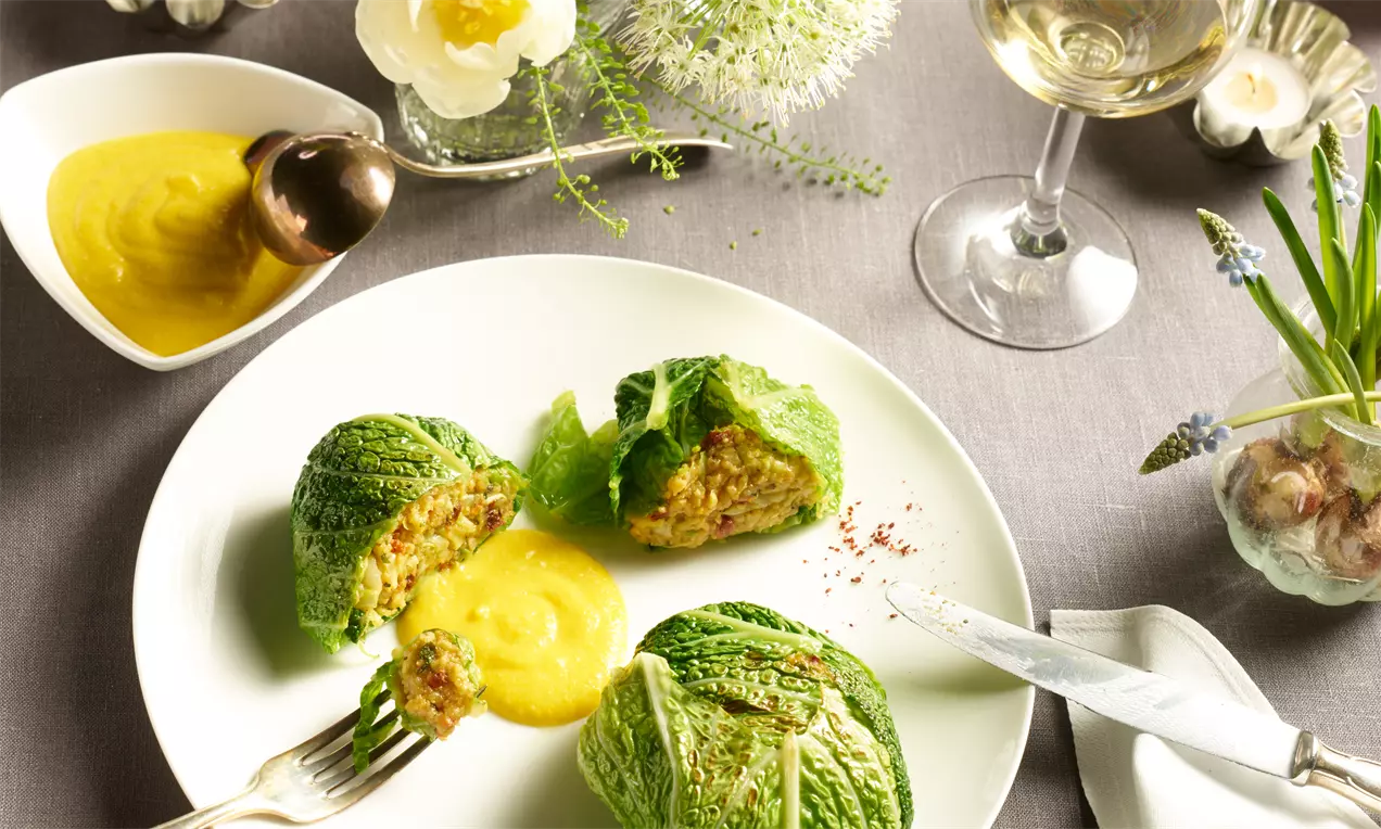 Grün umhüllte Kohlköpfchen mit lockerer Couscous-Füllung, begleitet von gelber Sauce auf weißem Teller mit zartem Besteck