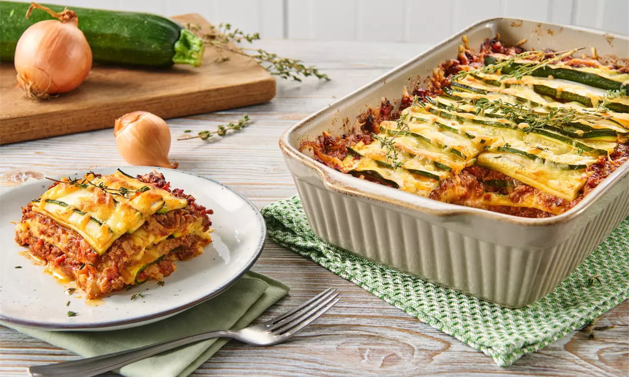 Zucchini-Lasagne in rechteckiger Ofenform mit goldbraun gebackenem Käse, grünen Zucchinistreifen und Kräutern, daneben ein angerichtetes Stück auf Teller