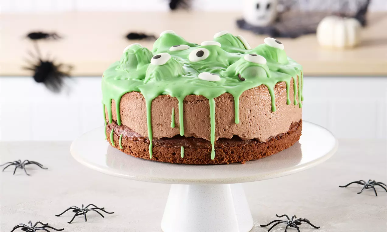 Schokoladen-Halloween-Torte mit grünem Zuckerguss, Augen-Verzierung und fluffiger Füllung auf weißer Tortenplatte, umgeben von Deko-Spinnen