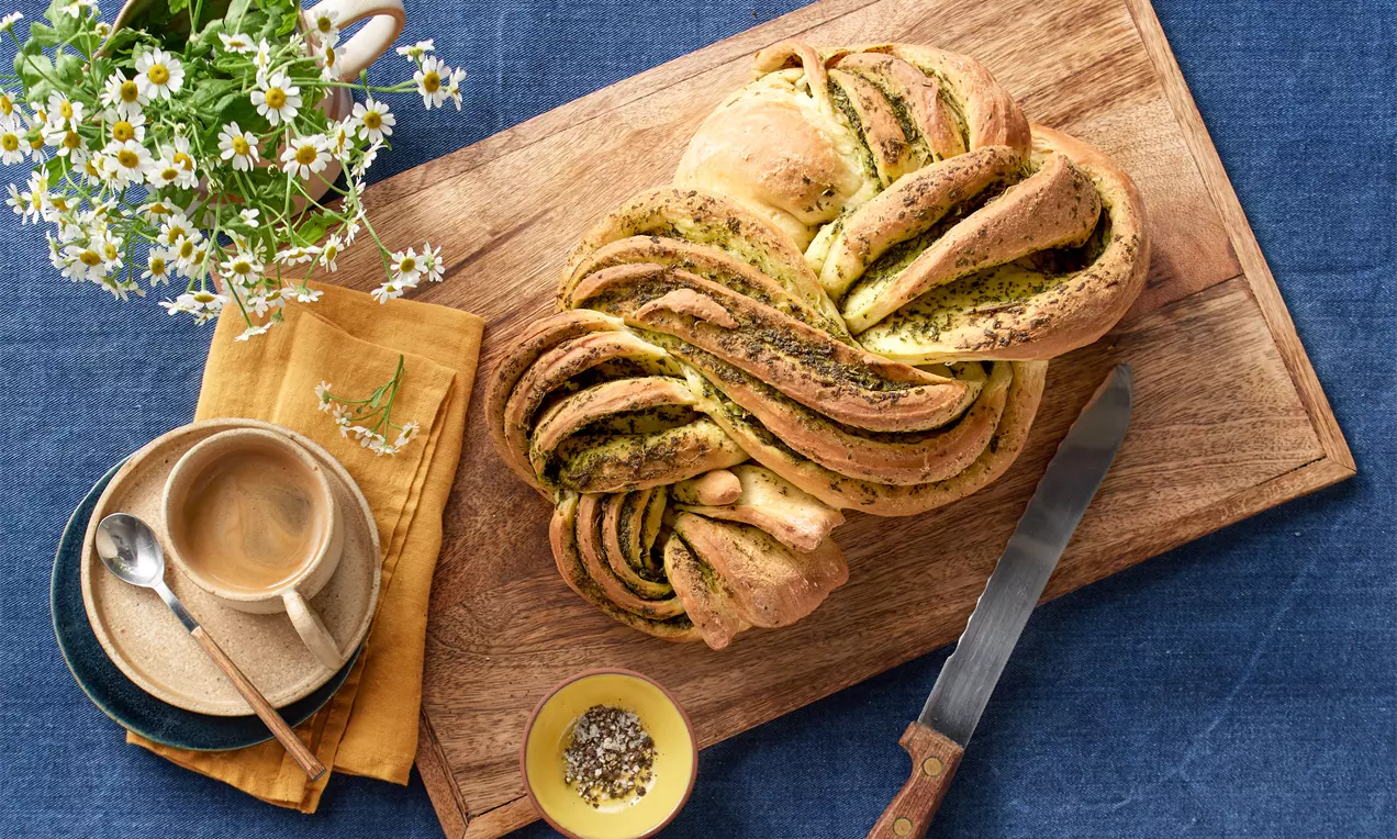 Goldbraunes, geflochtenes Rucola-Kräuterbrot auf einem Holzbrett, umgeben von einer Tasse Kaffee, Kamille und einem gelben Stofftuch