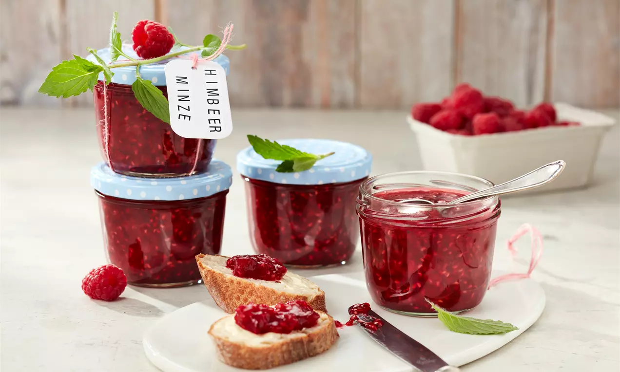 Himbeerrote Konfitüre mit Minzblättern in Gläsern mit blauem Deckel, aufgestrichen auf Brotscheiben auf weißem Teller mit frischen Himbeeren