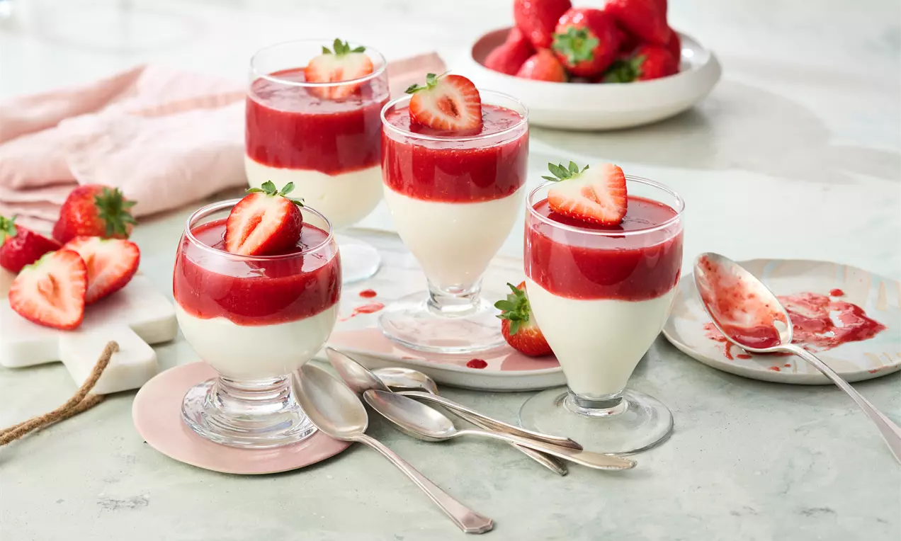 Puddingartige Creme mit glatter, weißer Textur, bedeckt von roter Erdbeersoße und halbierten Erdbeeren in gläsernen Dessertbechern