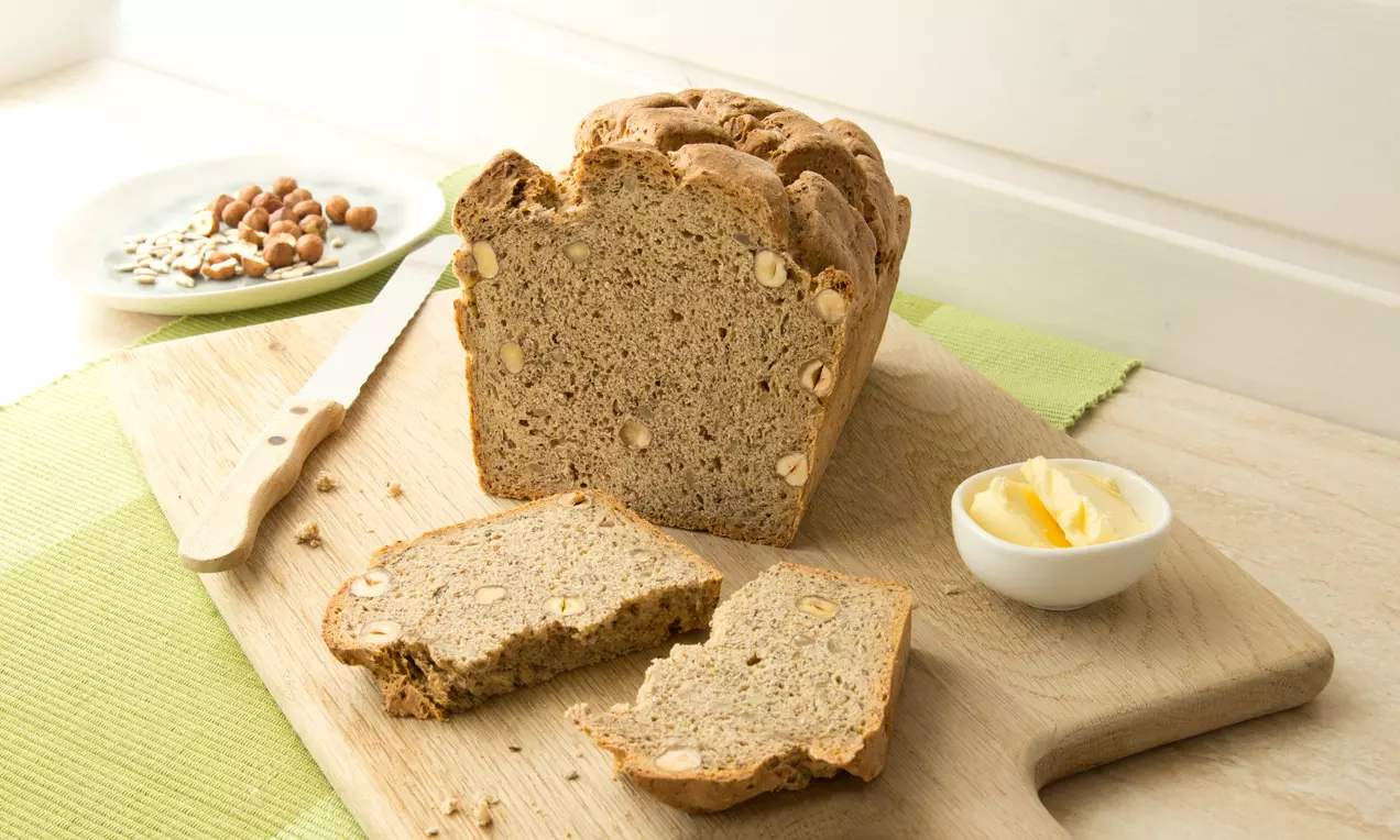 Dunkles Körner-Nuss-Brot mit goldbrauner Kruste und sichtbaren Nussstücken auf Holzbrett, daneben Butter, Messer und Kerne auf Teller
