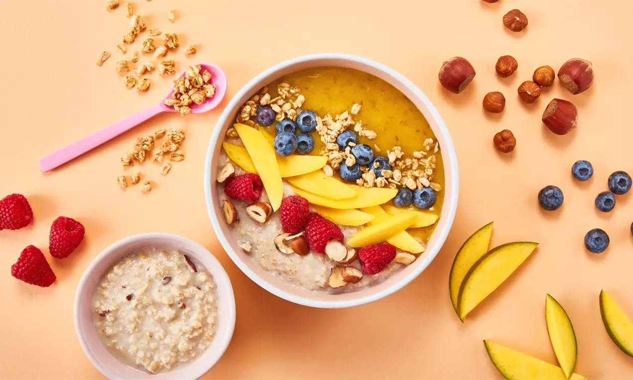 Bunte Frühstücksbowl mit gelber Mango, roten Himbeeren, blauen Heidelbeeren, Haselnüssen und Granola dekorativ auf cremiger Basis