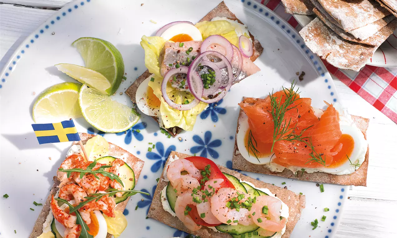 Knuspriges Knäckebrot mit bunten Belägen aus Lachs, Ei, Garnelen und Gemüse auf einem weißen Teller mit blauem Muster und Zitronenspalten